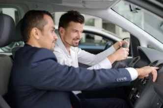 Vendedor de terno apontando para o painel de um carro enquanto o cliente ao volante observa, em test drive de carro