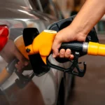 Mão abastecendo carro com bico de combustível enquanto avalia escolha entre álcool ou gasolina