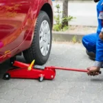 Profissional utilizando um macaco de carro para erguer um veículo vermelho durante manutenção ou troca de pneu.