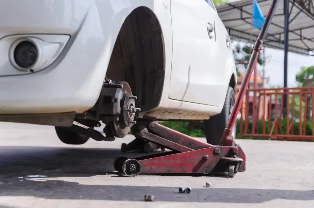 Macaco jacaré elevando um carro para manutenção, com a roda removida e peças apoiadas no chão durante o reparo automotivo.