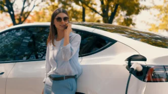 Carro híbrido elétrico estacionado enquanto mulher usa celular, com veículo conectado à tomada de recarga em ambiente externo.