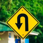 Placa de trânsito amarela em close-up indicando retorno em U, com fundo de vegetação; imagem que exemplifica placas de trânsito e sinalização viária para motoristas.